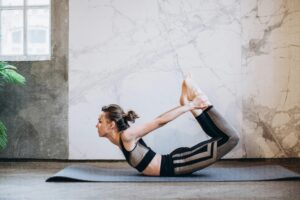 Person performing Dhanurasana, also known as Bow Pose, on a yoga mat in a serene indoor setting with a modern, minimalist background and green plants nearby.