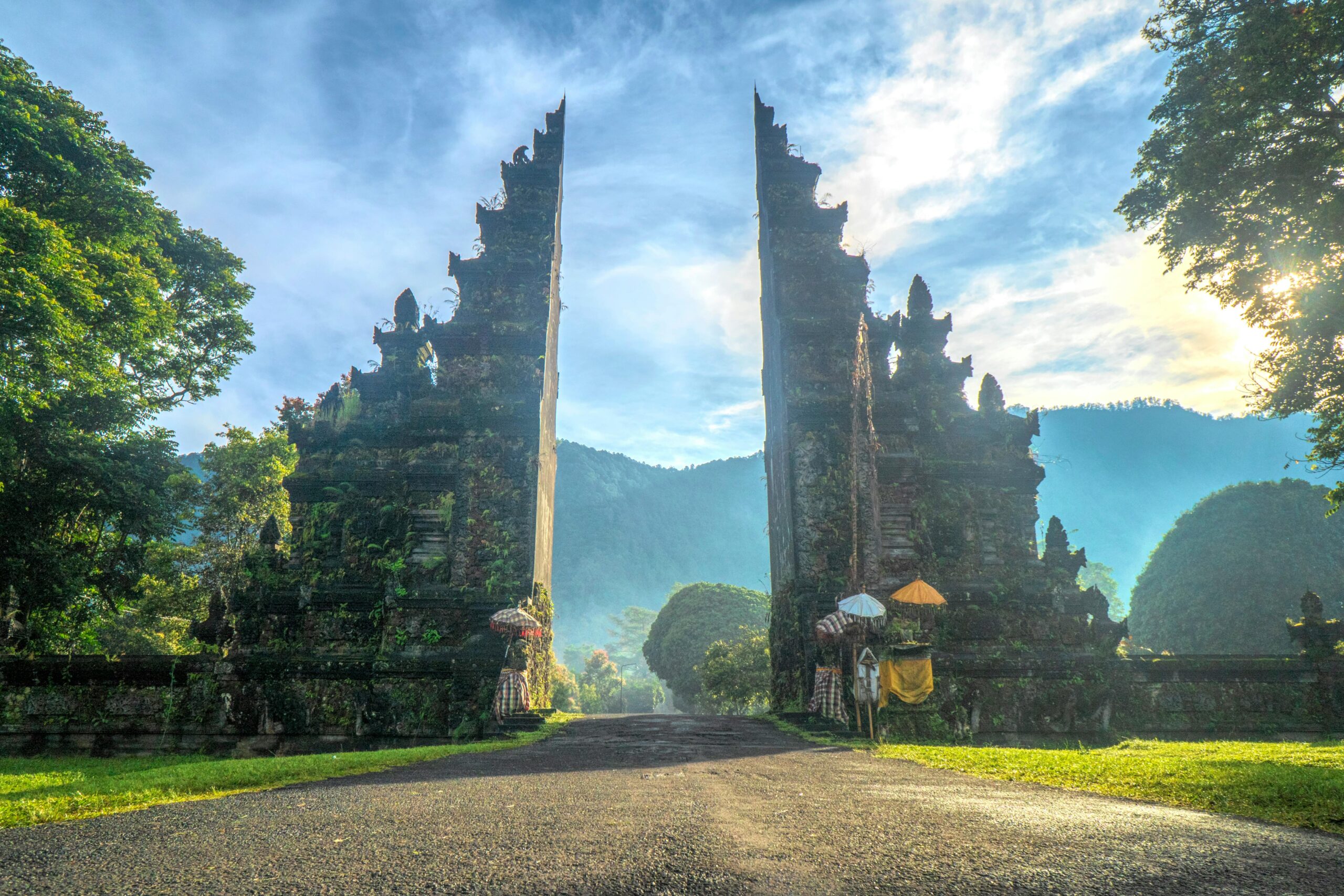 A scenic view of a traditional Balinese temple gate surrounded by lush greenery and mountains. The gate is partially covered in vines and moss, with decorative umbrellas in front. The sky is bright with a hint of sunlight, creating a serene and tranquil atmosphere