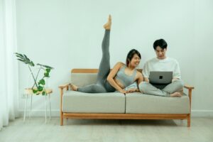 The image shows a woman in yoga attire performing a stretch on a couch while a man sits next to her, working on a laptop. The atmosphere is casual and relaxed, with a simple, modern interior design. A potted plant sits on a stand next to the couch, adding a natural touch to the scene. The image captures the blend of fitness, flexibility, and remote work, which is representative of the lifestyle of many digital nomads who balance their work and well-being from anywhere, even in their living space.