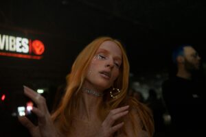 Close-up of a woman with glitter makeup and long red hair, wearing a choker and earrings, dancing in a vibrant nightclub setting with blurred background lights and people.